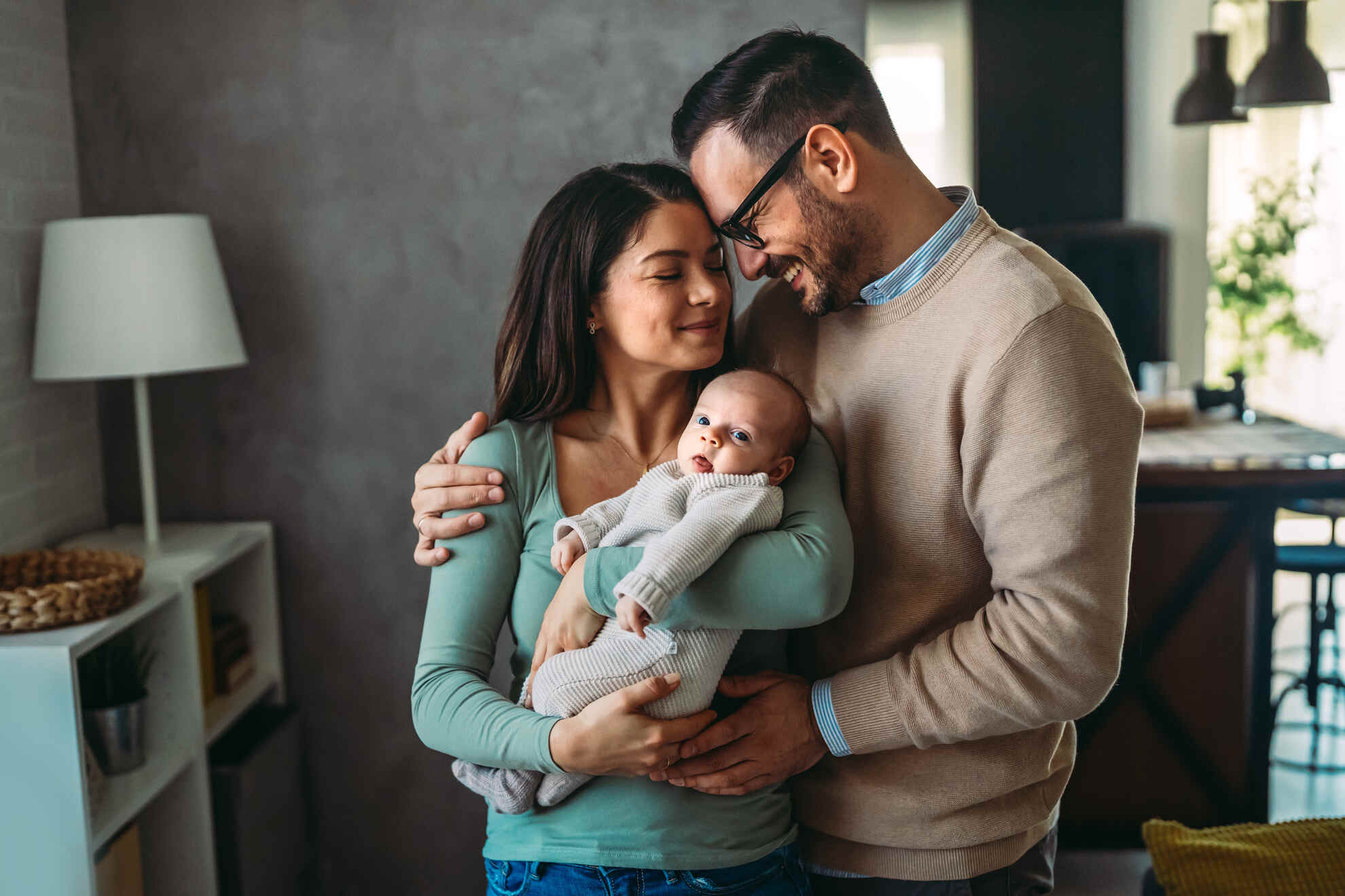 A smiling mother and father embrace while holding their infant baby in a cozy living room.