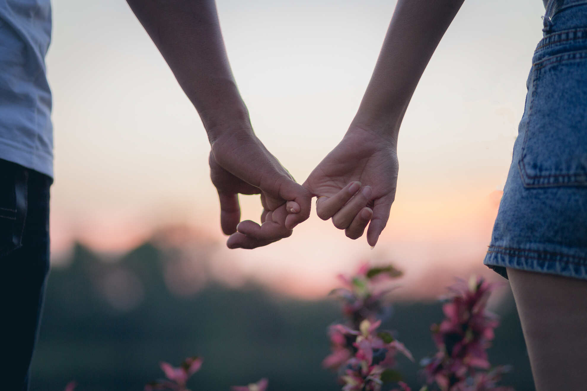 A close-up of two people's hands linked by their pinky fingers against a sunset backdrop.
