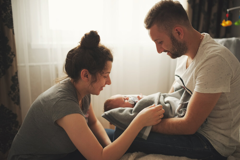 A mother and father sit close together, smiling down at their newborn baby wrapped in a gray blanket.