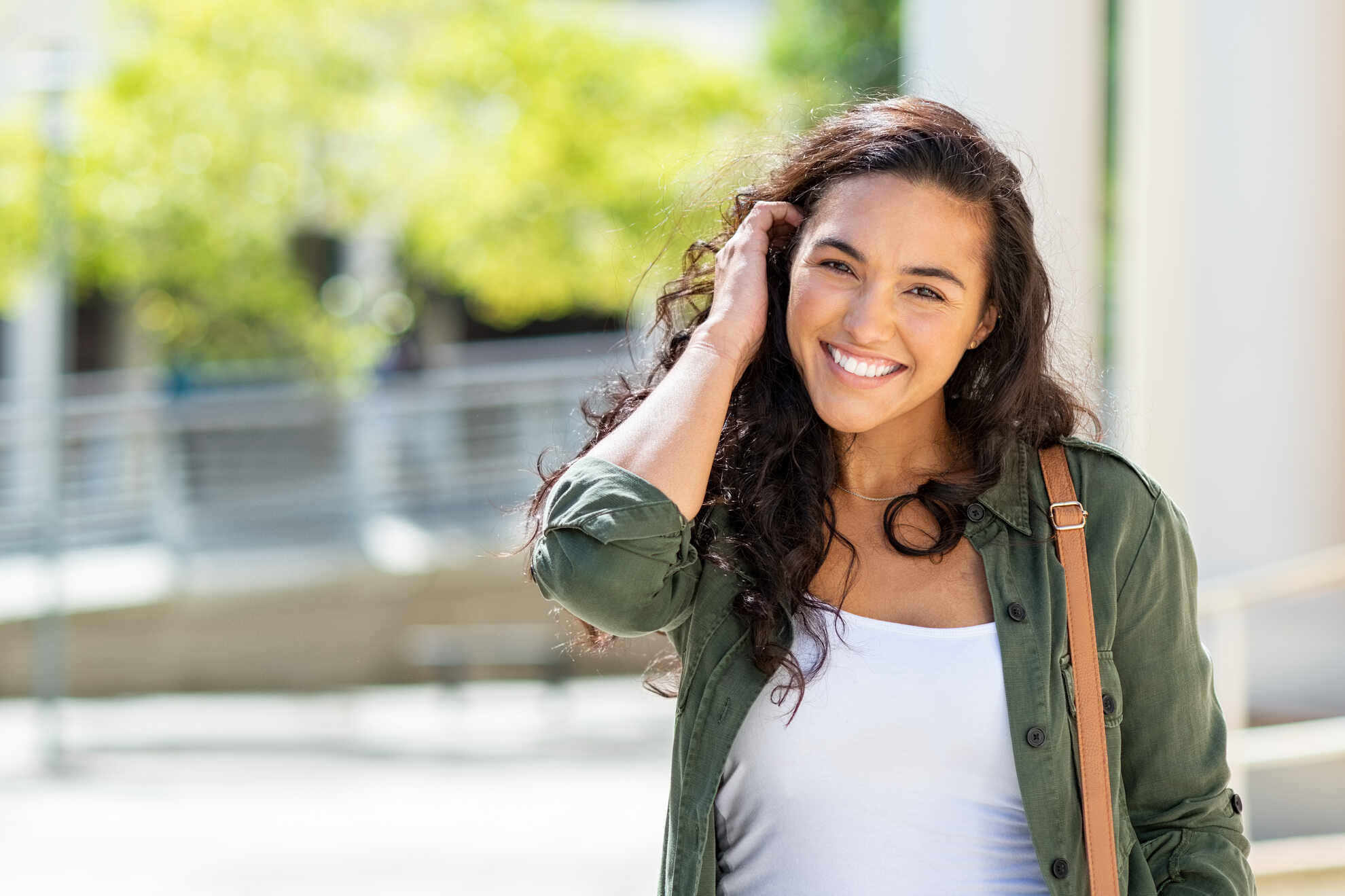 A smiling woman with long wavy dark hair wears an olive green shirt and white top outdoors.