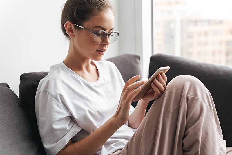 Woman wearing glasses sitting on a sofa and looking at a smartphone.