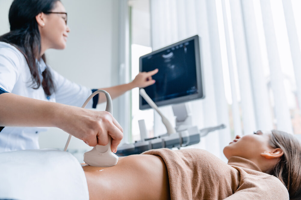 Young woman receiving an ultrasound from a female medical staff member wearing a white medical coat.