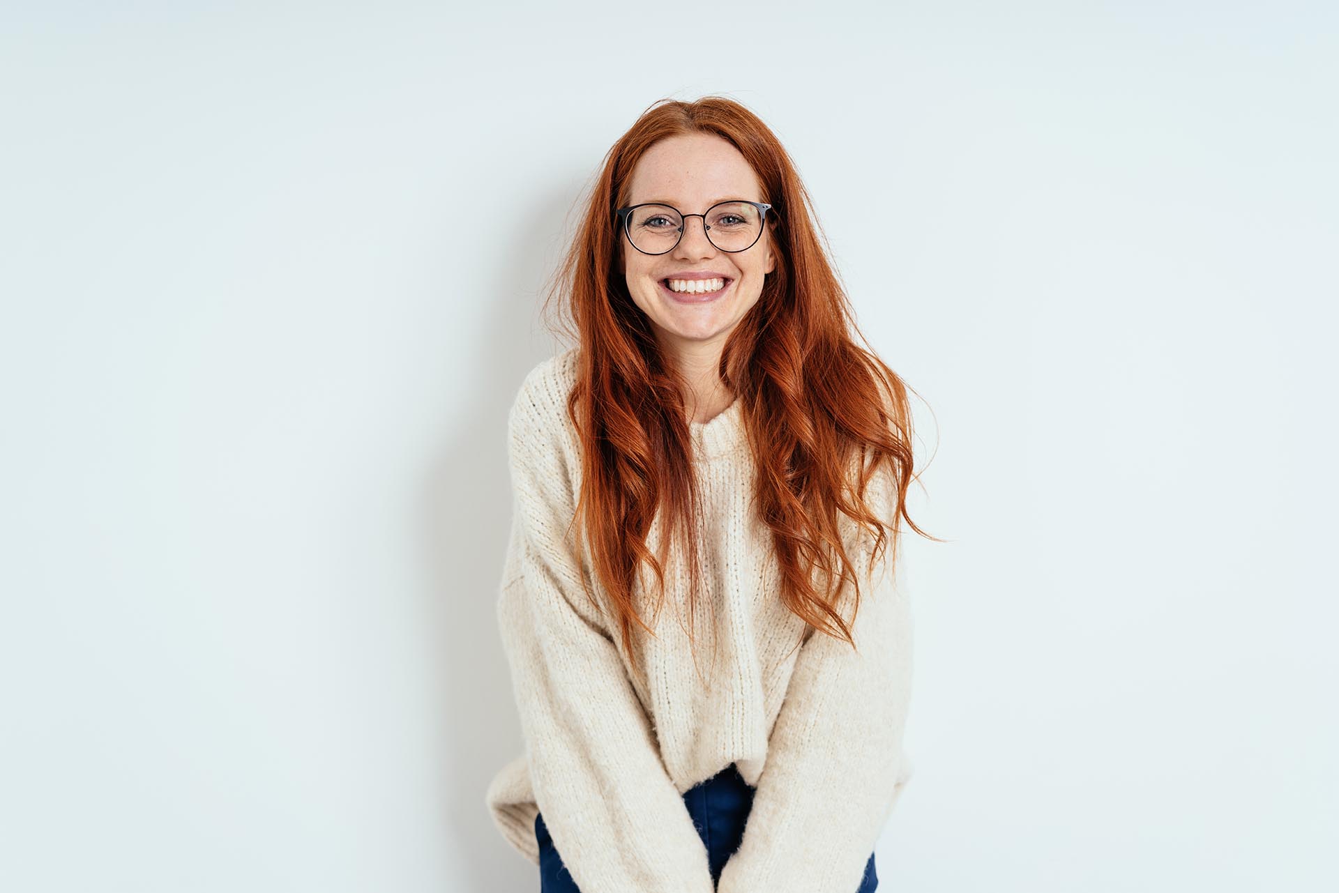 Happy young woman with red hair and glasses smiling at the camera with her back against a pure white backdrop.