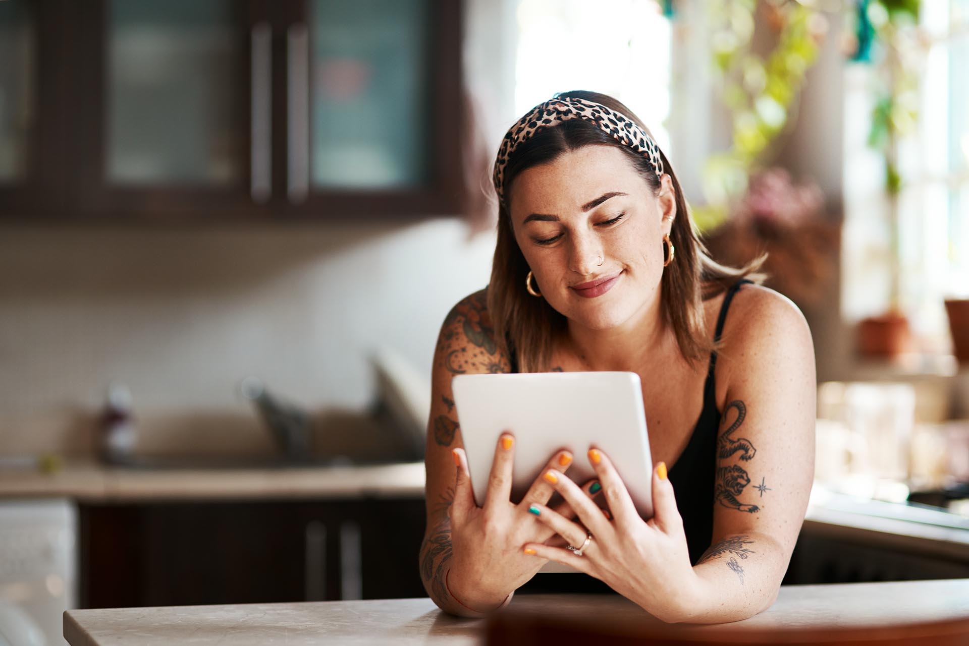 Tattooed woman wearing a leopard print headband smiles while looking at a tablet in a kitchen.