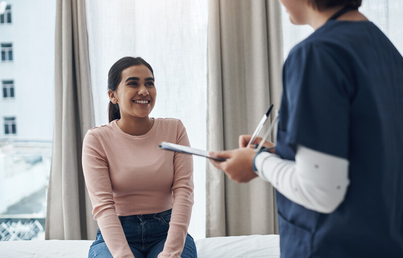 A smiling woman sitting on an exam table looks up at a healthcare worker holding a clipboard and pen.