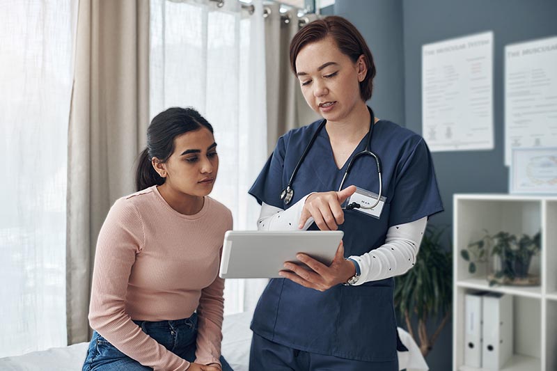 A female healthcare provider in blue scrubs shows information on a tablet to a young woman sitting in a clinic room.