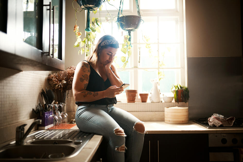 A woman with tattoos sits on a kitchen counter near a bright window while looking at her smartphone.