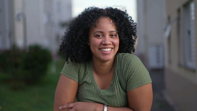 Smiling woman with curly black hair wearing a green ribbed shirt outdoors.