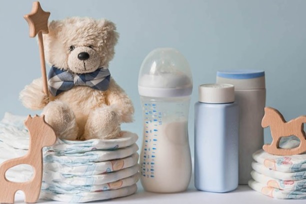 Teddy bear on stacked diapers next to a baby bottle, blue toiletries, and wooden toys against a light blue background.
