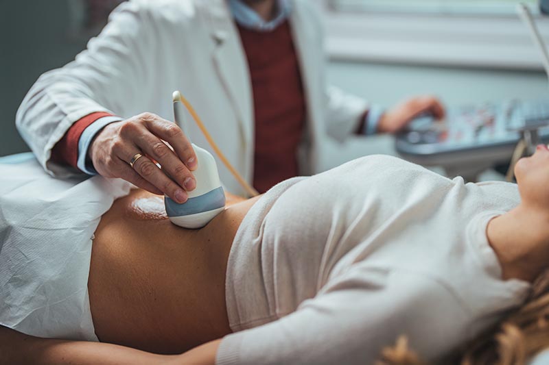 A doctor in a white coat performs an abdominal ultrasound on a woman lying on an examination table.