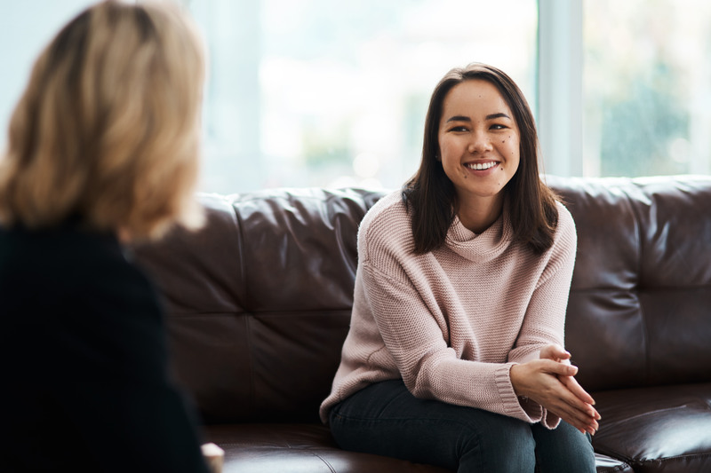 A smiling young woman in a pink sweater sits on a leather couch, engaged in conversation with an out-of-focus companion.