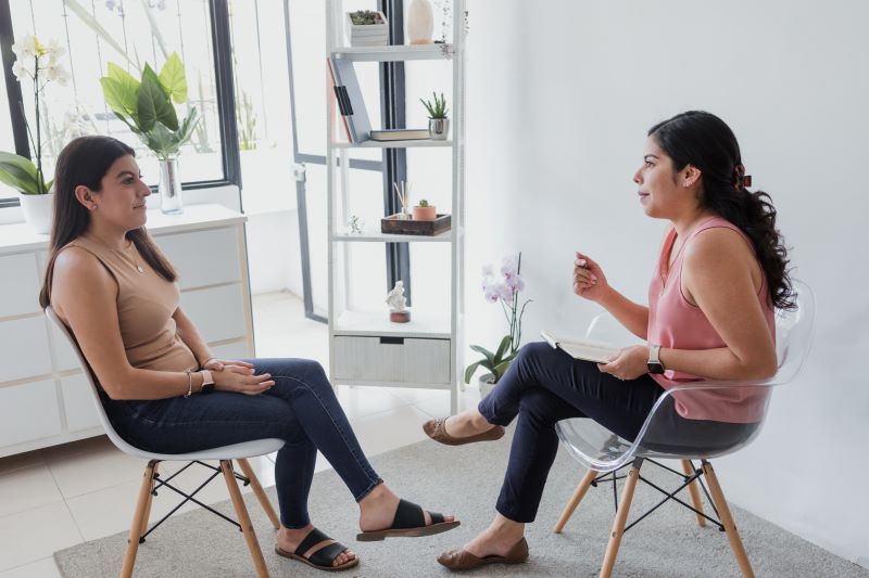 Two women sit facing each other in chairs during a consultation, one taking notes while the other listens.