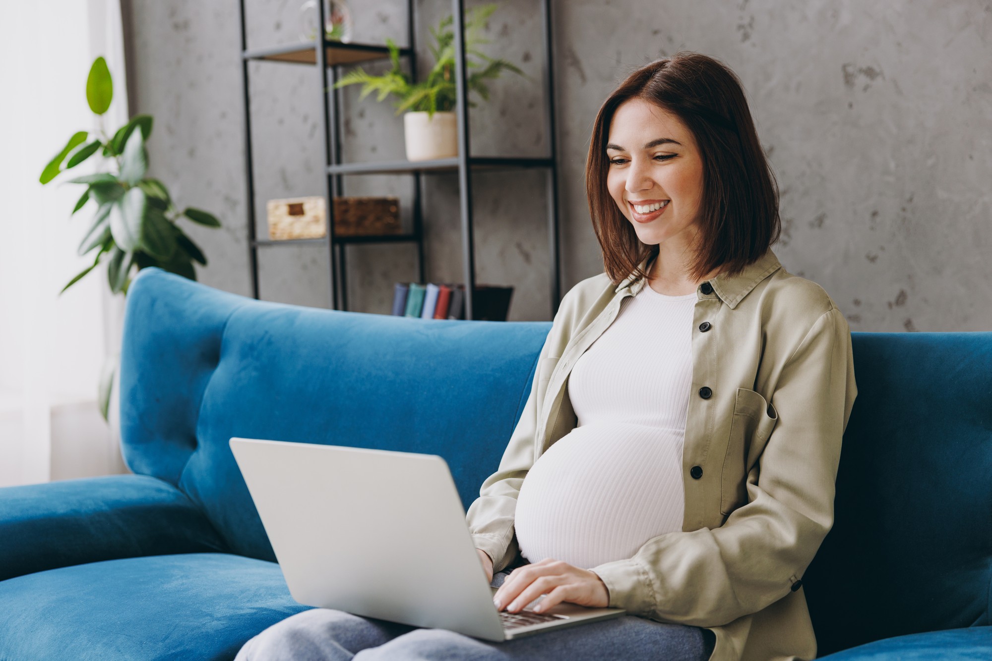 Smiling pregnant woman sitting on a blue sofa while using a laptop computer.