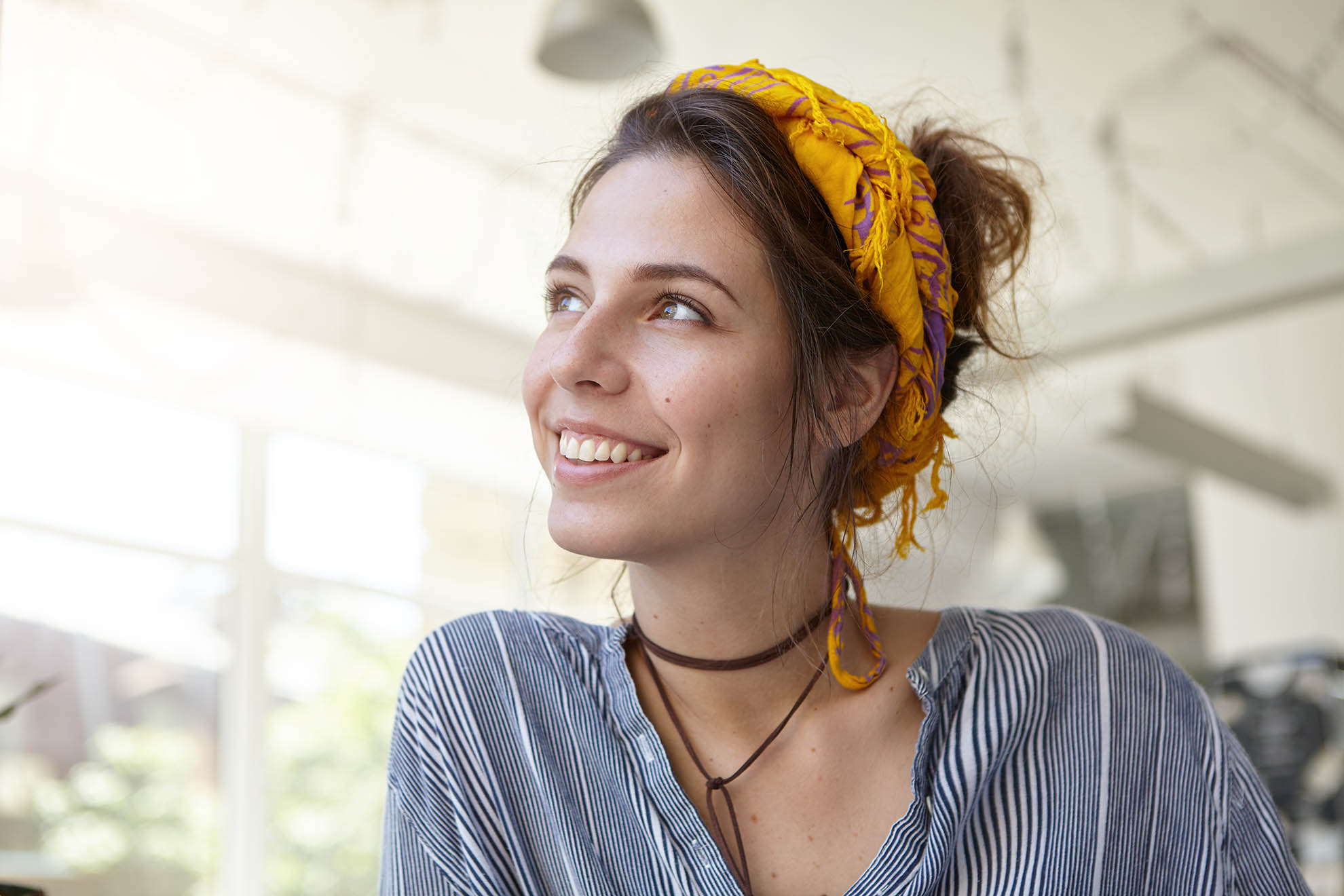 Sideways portrait of beautiful Caucasian woman with headband on head and casual shirt looking with smile aside enjoying bright light and warm sun. Office worker having rest indoor dreaming about rest Young woman in a plaid shirt smiles while discussing paperwork at a desk in an office or library.